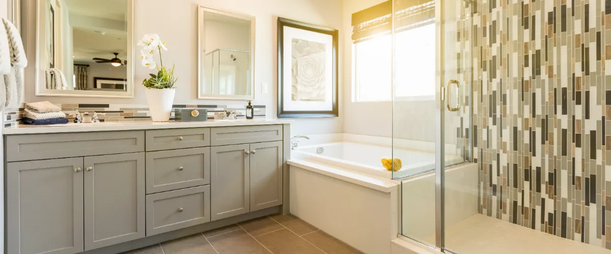 A bright, modern bathroom with a glass shower, freestanding tub, gray cabinetry, and decorative tile accents. Sunlight streams in through the window