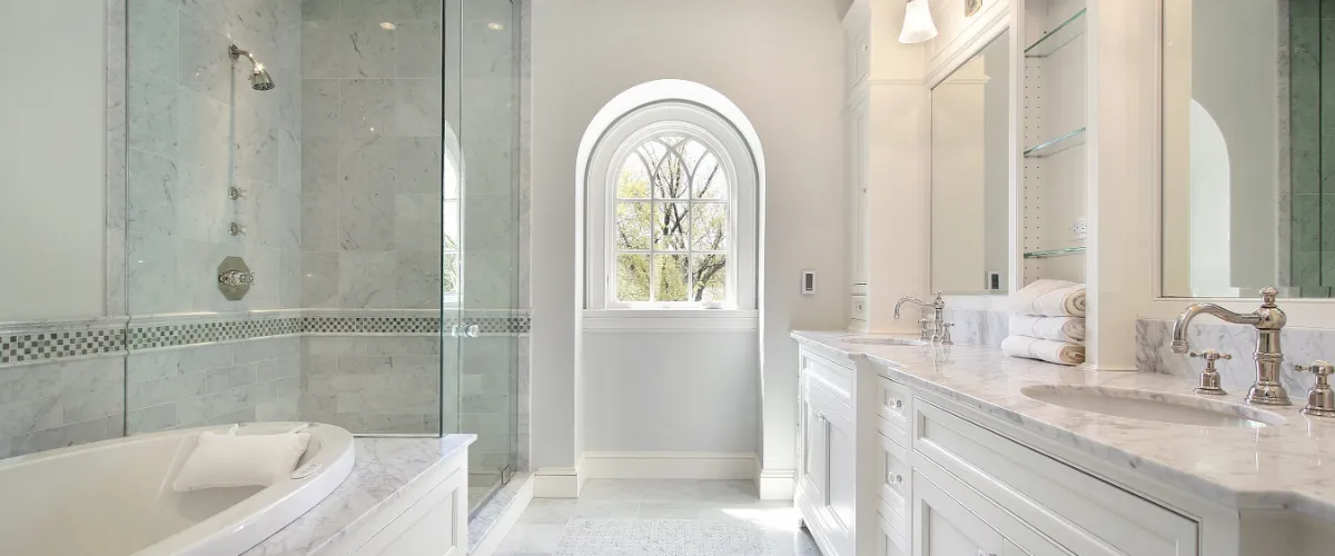 Spacious modern bathroom featuring a glass shower, freestanding tub, double vanity, and large arched window overlooking greenery