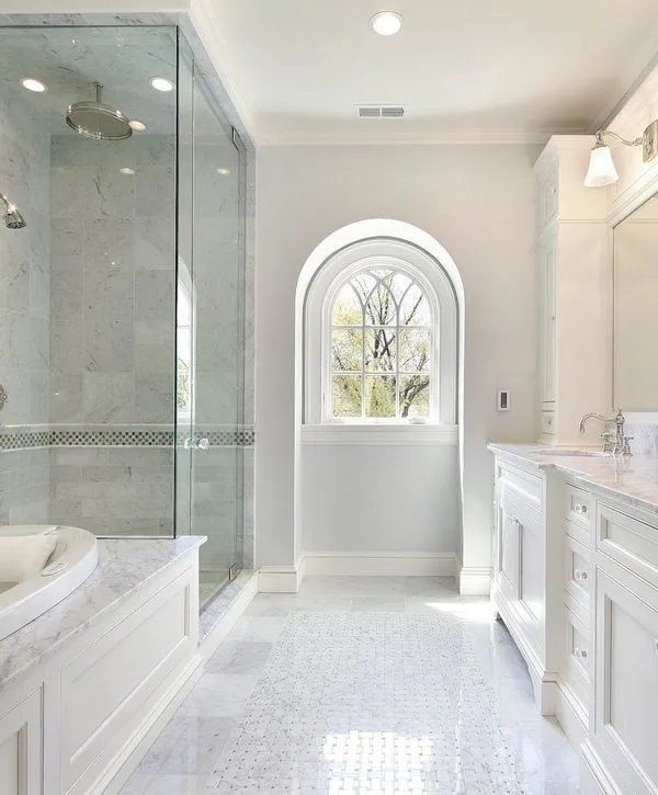Elegant master bathroom with white cabinetry, marble countertops and floor, a spacious glass shower, and an ornate arched window letting in natural light