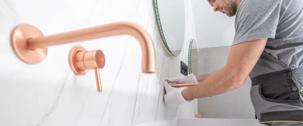 a worker installing a modern wall mounted, rose gold or copper toned faucet in a bathroom with white marble tiles