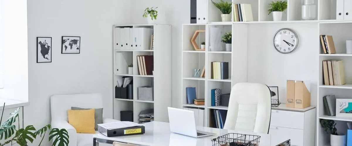 A bright, clean home office created in a basement space, featuring white walls, extensive white shelving units filled with files and books, and a comfortable white ergonomic chair.
