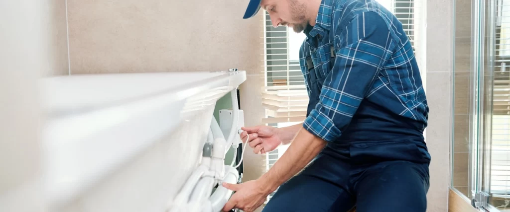A certified technician or plumber, wearing a blue plaid shirt and cap, is bent over connecting wires and plumbing components underneath the side panel of a white bathtub during installation.
