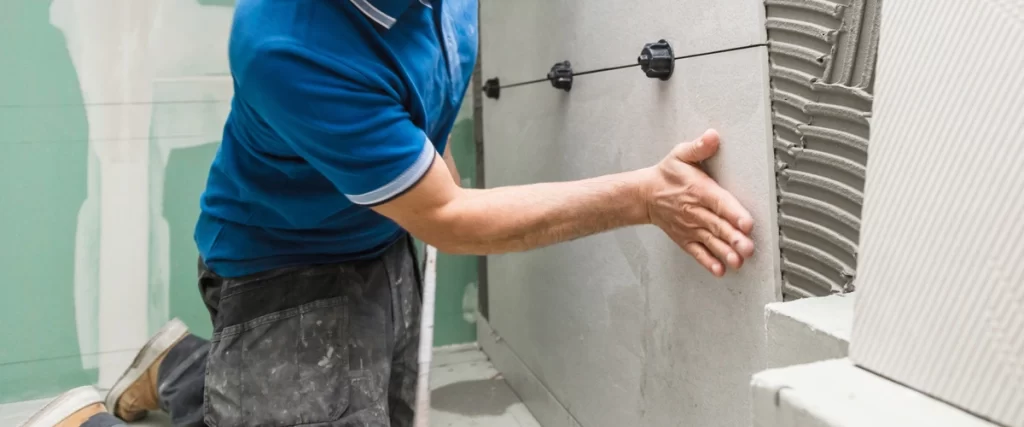 A contractor, kneeling down, is pressing a large cement or drywall panel onto a prepared wall, using black tile leveling clips for precise installation during a bathroom remodeling project.