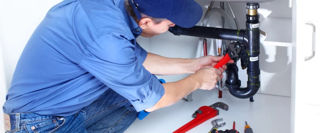 A licensed plumber in a blue shirt and cap crouches under a white bathroom sink, using a red-handled wrench to tighten or repair the P-trap and drain piping.