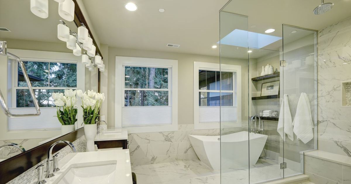 A luxurious, light-filled master bathroom remodeling project featuring white marble tile, a standalone geometric soaking tub, a seamless glass shower with a skylight, and a double vanity with striking light fixtures.