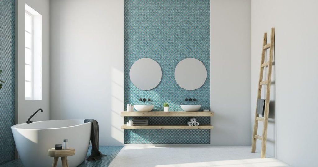 A beautifully remodeled, bright, modern bathroom featuring a freestanding white bathtub, two circular vessel sinks on a wooden shelf against a blue mosaic accent wall, and a rustic wooden ladder for towels.
