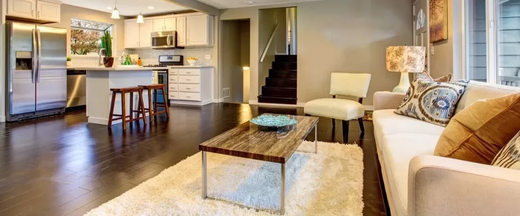 A spacious open-concept basement featuring a modern kitchen with stainless steel appliances, white cabinetry, an island with bar stools, dark hardwood floors, and a cozy living area with a rug, sofa, accent chair, and decorative pillows.