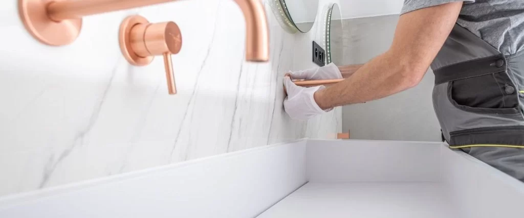 A plumber wearing white gloves carefully installs a sleek rose gold wall-mounted faucet and handles onto a white marble countertop and backsplash above a white vanity sink.