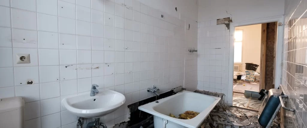 A partially gutted bathroom showing white tiled walls, a small sink, and a bathtub filled with debris during the bathroom demolition process.