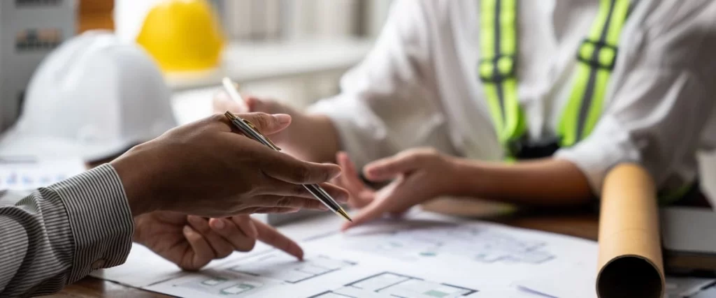 Two construction professionals in safety gear pointing at architectural blueprints while planning the layout for new bathroom floor tiles.