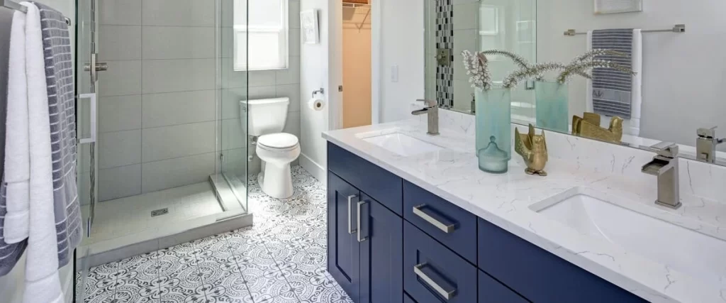 A bright bathroom interior showcasing a navy blue double vanity, quartz countertops, and intricate black and white patterned mosaic floor tiles.