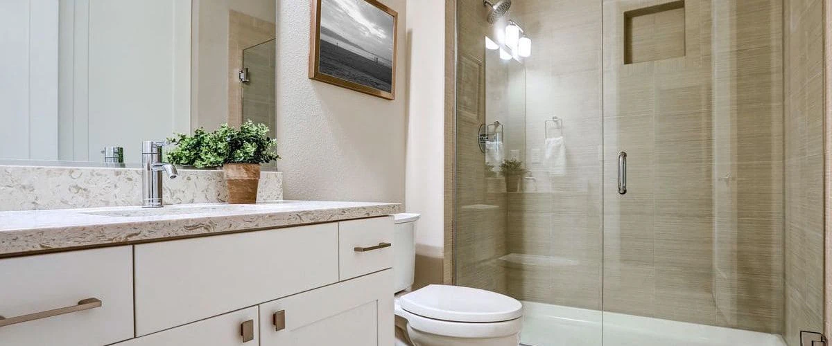 A bright contemporary bathroom showcasing a glass-enclosed walk-in shower, a white vanity with a marble countertop, a small potted plant, and light beige wall tiling.