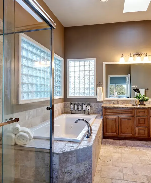 A spacious master bathroom remodeling project in Aberdeen, NJ, featuring a large white soaking tub with stone tile surround, glass block windows for natural light, and a warm wood double vanity with granite countertops.