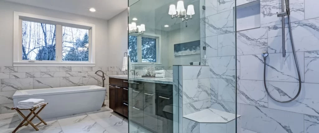 A spacious master bathroom featuring large format white marble wall tiles, a glass-enclosed walk-in shower with a built-in bench, and a modern freestanding bathtub.