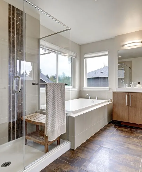 A bright, modern bathroom remodeling in Rumson, NJ featuring a glass-enclosed walk-in shower with vertical mosaic tile, a wooden spa bench, and a large white soaking tub under a window.