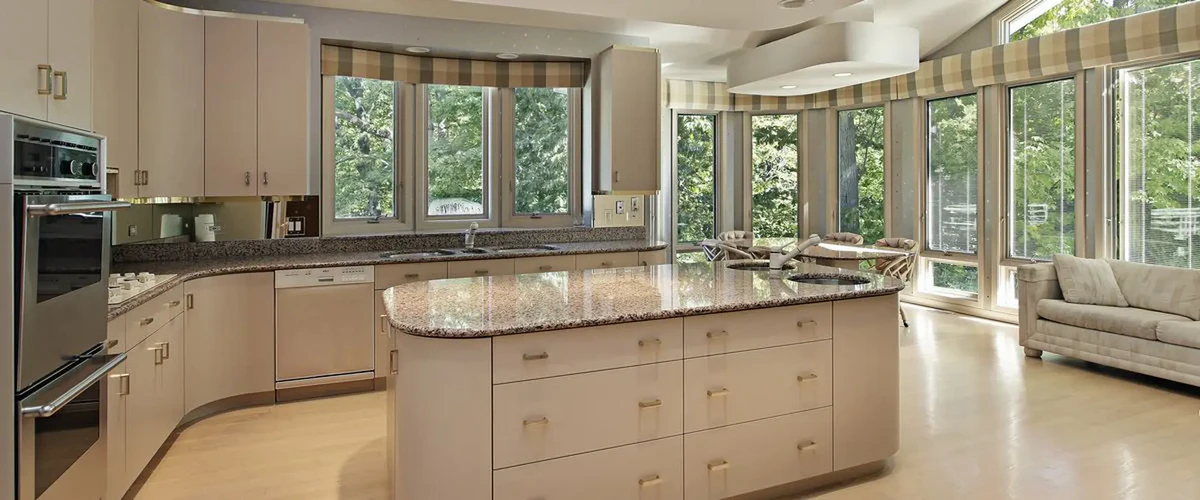 A bright modern kitchen design with a curved granite-topped island, neutral cabinets, and floor-to-ceiling windows overlooking a lush green landscape.