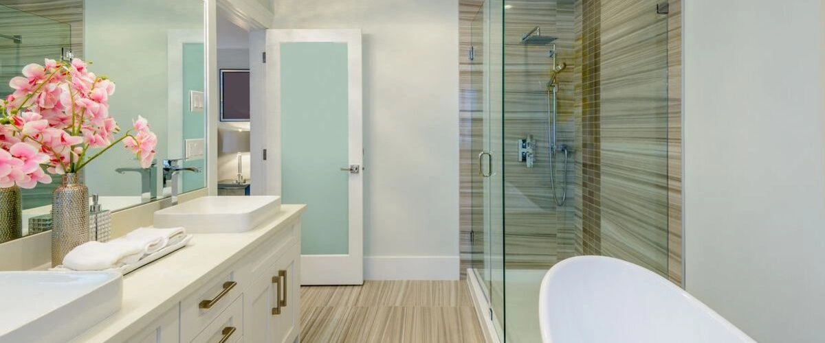 A bright modern bathroom design showcasing a glass-enclosed walk-in shower, white cabinetry with gold hardware, and pink orchids on the counter.