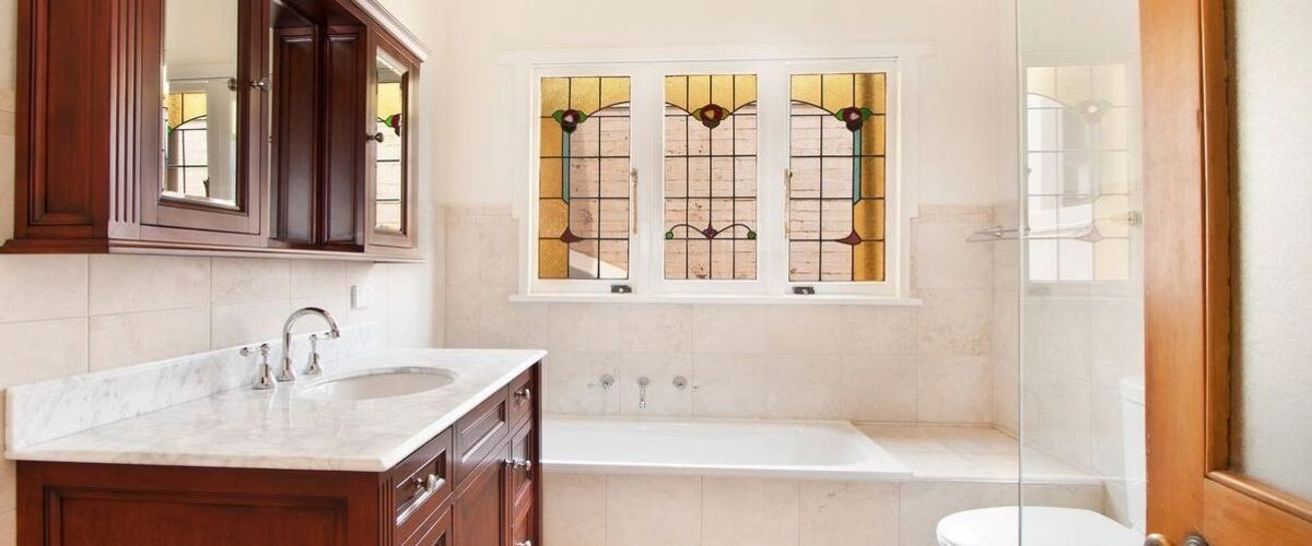 A classic vintage bathroom featuring an ornate stained glass window, a dark wood vanity with a white marble countertop, and a built-in bathtub.