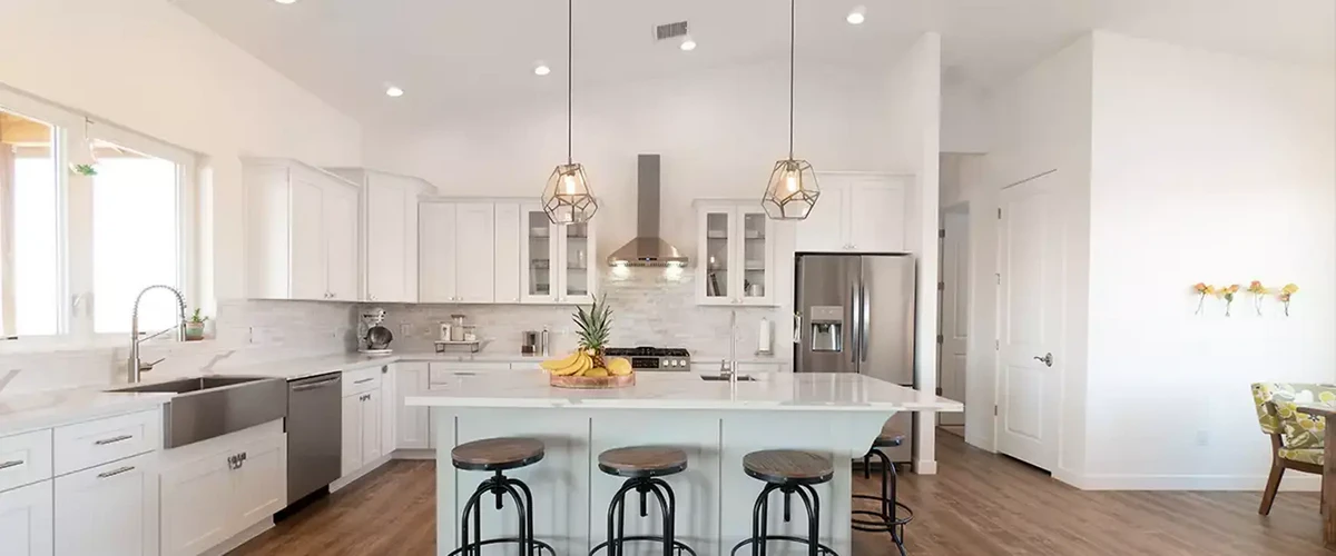 Bright and airy kitchen featuring white cabinetry, vaulted ceilings, cage pendant lights, and a large breakfast bar with wooden stools.