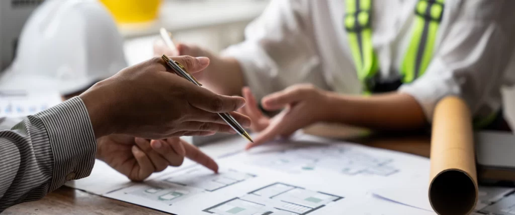 Close up of contractors in safety gear pointing at house floor plans on a wooden table to ensure compliance with local building codes.