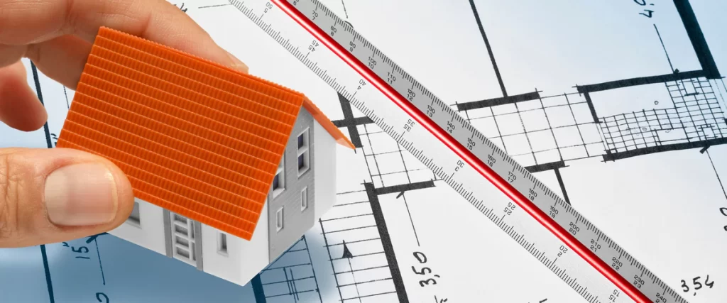 A hand holding a small house model over architectural floor plans and a red scale ruler, representing the planning and permit process for a kitchen renovation.