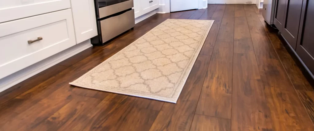 A perspective view of a dark wood-textured laminate kitchen floor featuring a light beige trellis pattern runner rug between white and dark cabinetry.