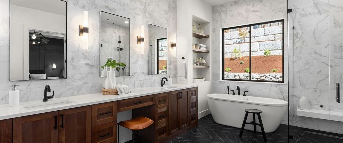 A luxury master bathroom featuring a dark wood double vanity, white marble wall tiles, a freestanding soaking tub, and black herringbone floor tiles near a large window.