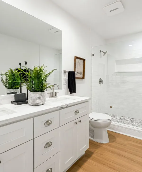 A bright, modern bathroom remodeling project in Port Monmouth, NJ featuring a white shaker vanity, marble countertops, a potted fern, and a walk-in shower with pebble tile flooring.