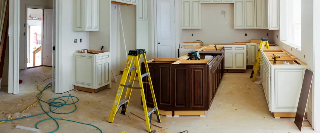 A kitchen remodeling scene featuring a dark wood center island, white upper cabinets, and yellow stepladders with construction tools and blue power cords on the floor.