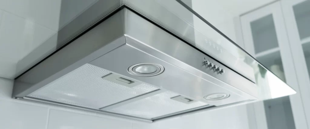 A close-up under-view of a modern stainless steel range hood featuring a glass canopy, mesh filters, and built-in LED lighting in a bright kitchen.