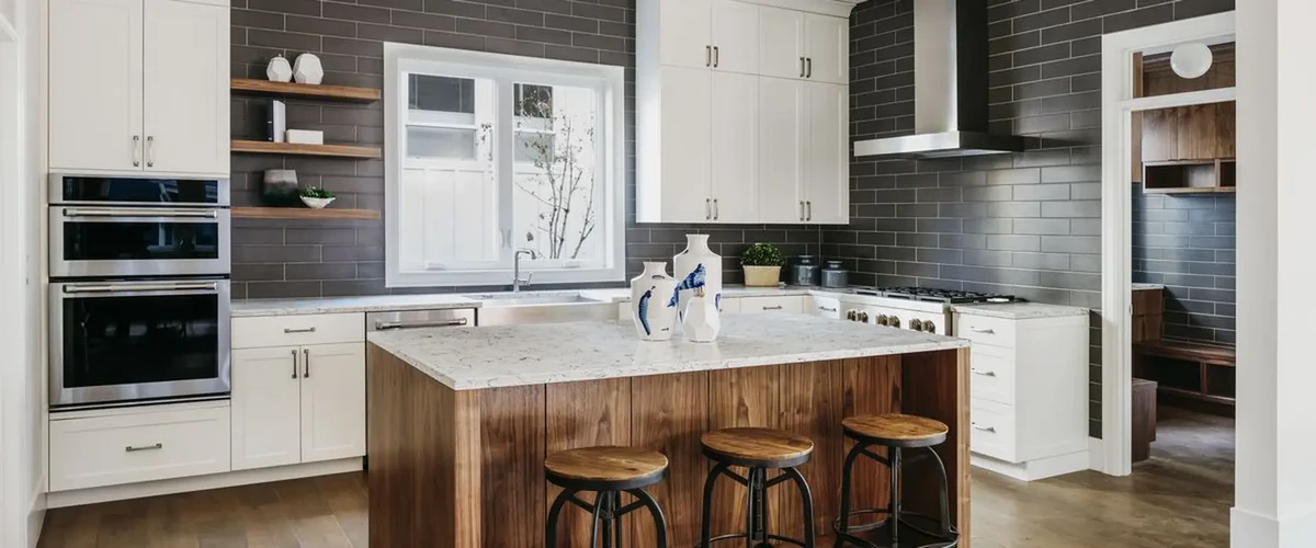 Wide view of a modern kitchen showcasing a large wood-paneled island, dark gray subway tile backsplash, white cabinetry, and stainless steel double wall ovens.