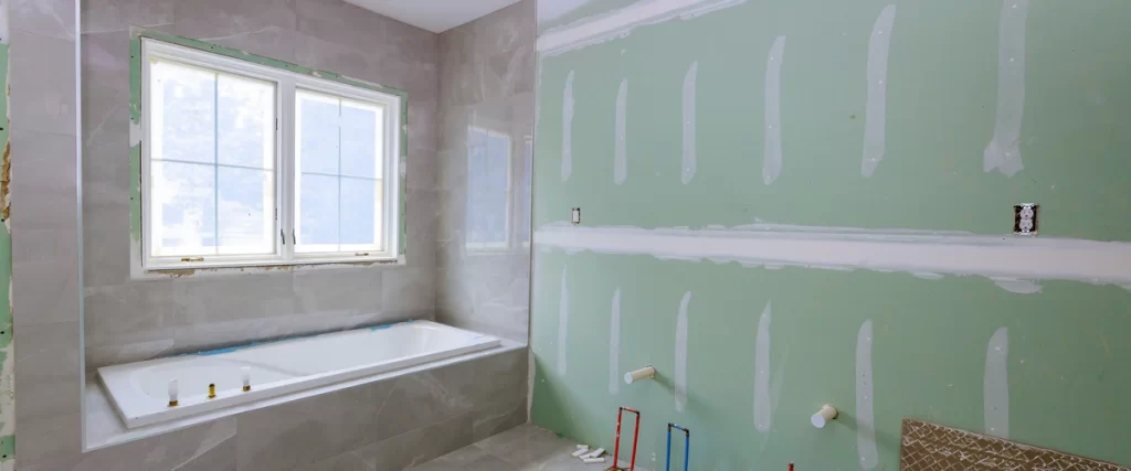 A bathroom in the middle of a room by room remodeling project, showing a newly installed white bathtub below a window, surrounded by green moisture-resistant drywall with taped seams and exposed plumbing pipes on the floor.