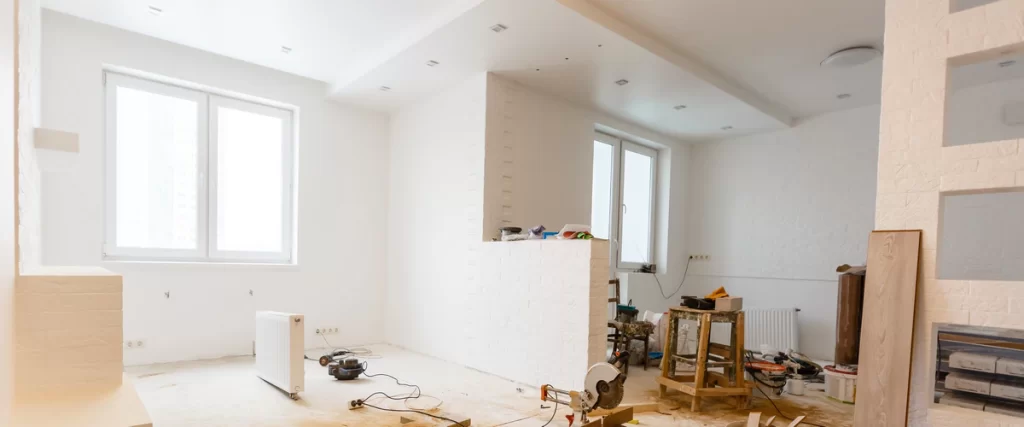 A bright, white room under renovation featuring a miter saw on the floor and a painted brick half-wall left over after removing load-bearing wall.