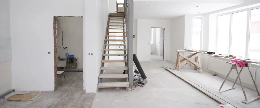 Interior view of a house undergoing whole home remodeling, featuring an unfinished wooden and metal staircase, exposed brick around a doorway, bare concrete floors, and construction sawhorses in a bright, gutted space.