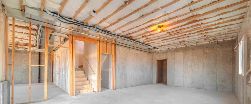 A wide view of an unfinished basement featuring bare concrete foundation walls, a wooden staircase, partial wood wall framing, and pink fiberglass insulation installed between the ceiling joists.