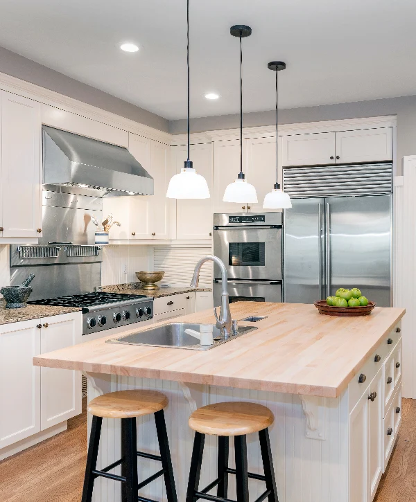 A bright kitchen space featuring a large light wood butcher block island with three white pendant lights, white cabinetry, and stainless steel appliances, completed as a custom home remodeling project in Manalapan Township, NJ.